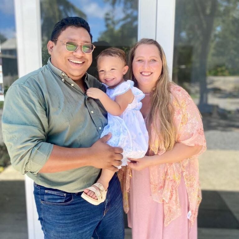 A joyful family of three from Roatan Honduras standing outdoors with a glass building and greenery in the background, smiling warmly at the camera.