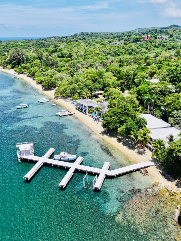 Aerial view of a tropical beach in Roatan, Honduras, with a marina and boats docked along the shoreline, lush green trees, and distant hills under a partly cloudy sky.