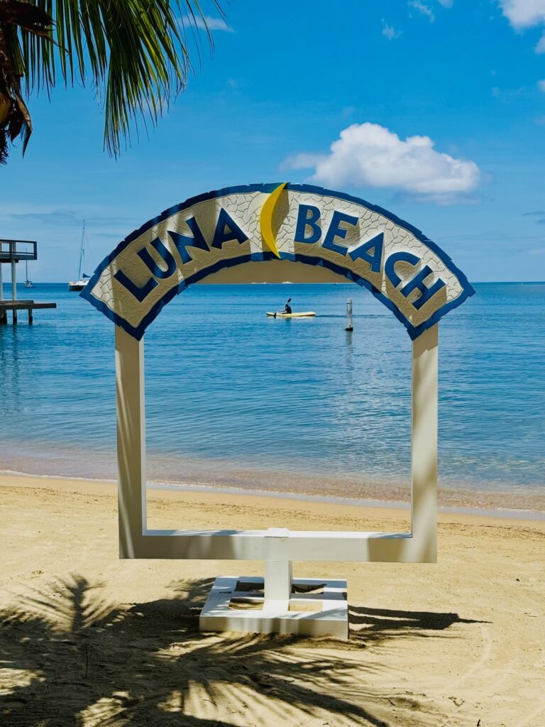 Roatan Beach sign with palm tree shadows and calm turquoise waters in the background of Honduras' Bay Islands.