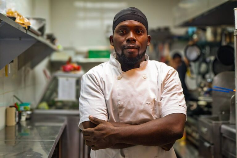 A professional chef in a white uniform and black headscarf stands with arms crossed inside a busy kitchen in Roatan, Bay Islands, Honduras, surrounded by cooking utensils and appliances.