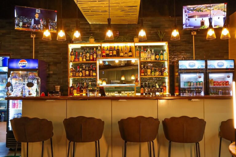 Interior view of a bar in Roatan, Honduras, featuring a well-stocked back bar with various liquor bottles, illuminated shelves, and modern pendant lighting, with televisions mounted on the dark wooden wall behind the bar.