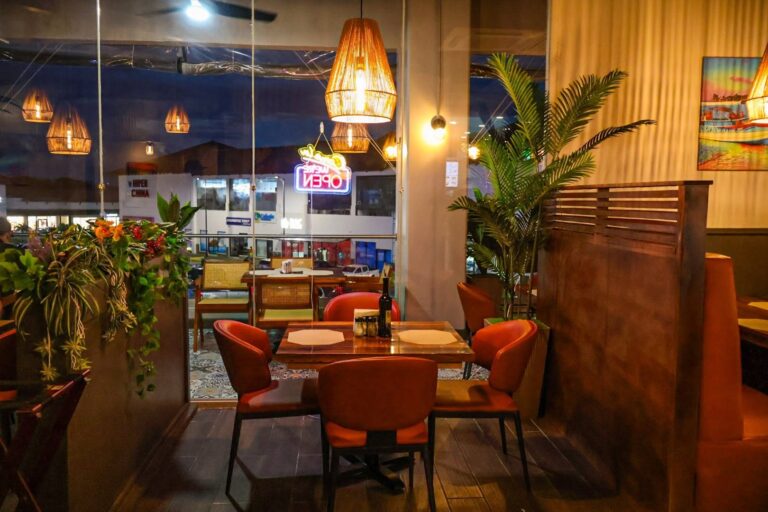 Indoor view of a cozy restaurant in Roatan, Honduras, illuminated by warm pendant lights, with large glass windows overlooking the lively street scene outside featuring a neon 'OPEN' sign and people dining at nearby establishments.
