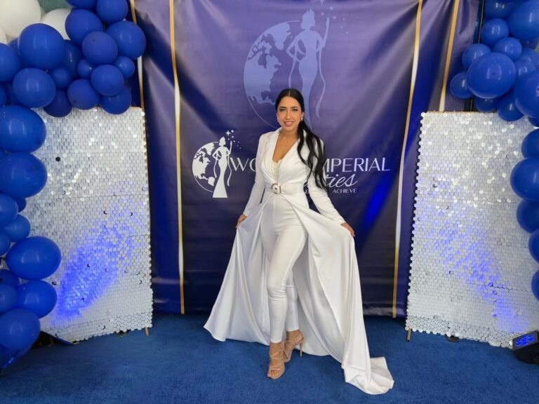 A woman dressed in a white outfit standing in front of a Roatan and Honduras themed backdrop decorated with blue balloons and white hexagonal panels.