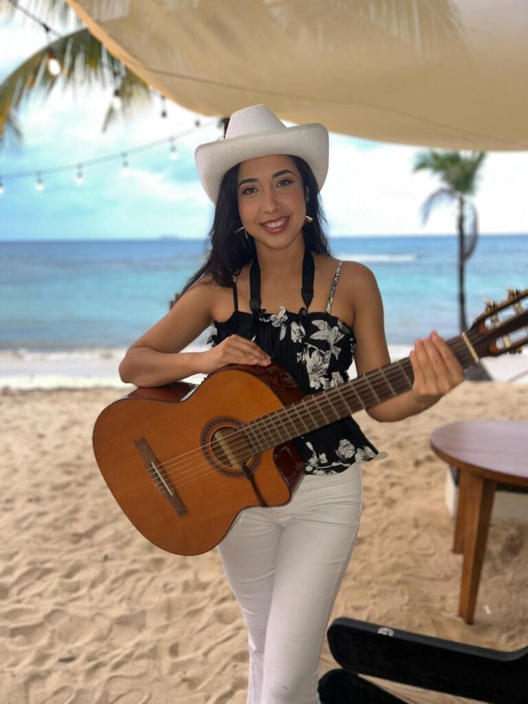 A woman with dark hair wearing a white cowboy hat and floral top playing an acoustic guitar on a sandy beach in Roatan Honduras, with palm trees and the ocean in the background.
