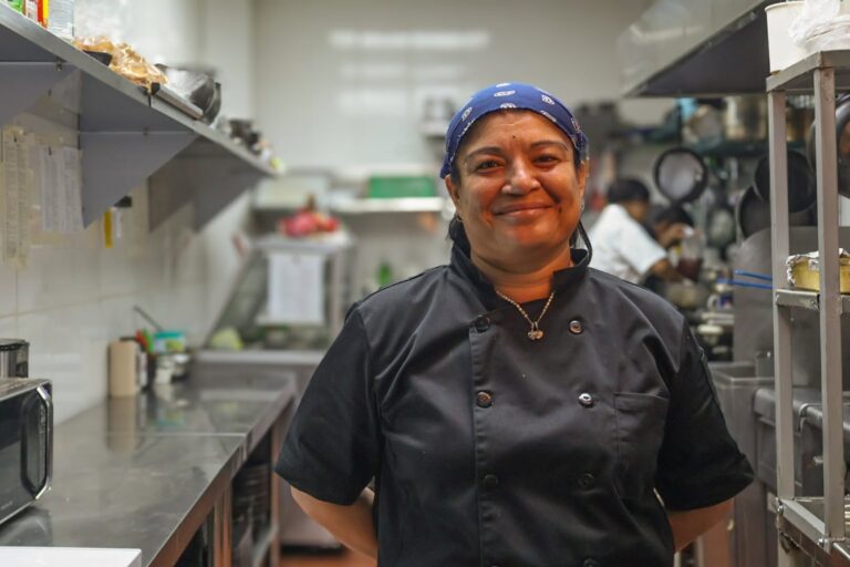 A smiling female chef in a black uniform and blue bandana stands in a professional kitchen on Roatan, Honduras, with stainless steel shelves and cooking equipment around her.