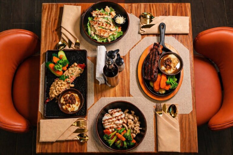 A top-down view of a wooden dining table set with four dishes, including grilled chicken and vegetables, a hearty meat stew with carrots and broccoli, a creamy pasta with vegetables, and a grilled steak with sides and sauce, surrounded by orange leather chairs in a restaurant on Roatan, Honduras.