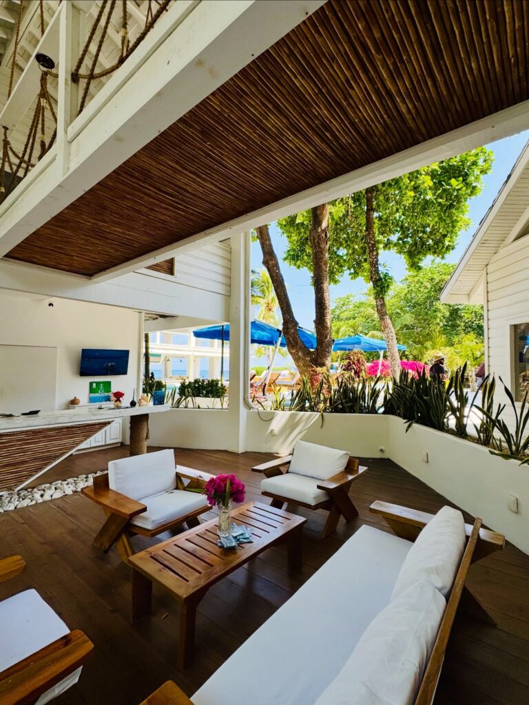 Outdoor seating area with wooden furniture, lush greenery, and vibrant pink flowers under a shaded wooden ceiling in Roatan, Honduras.