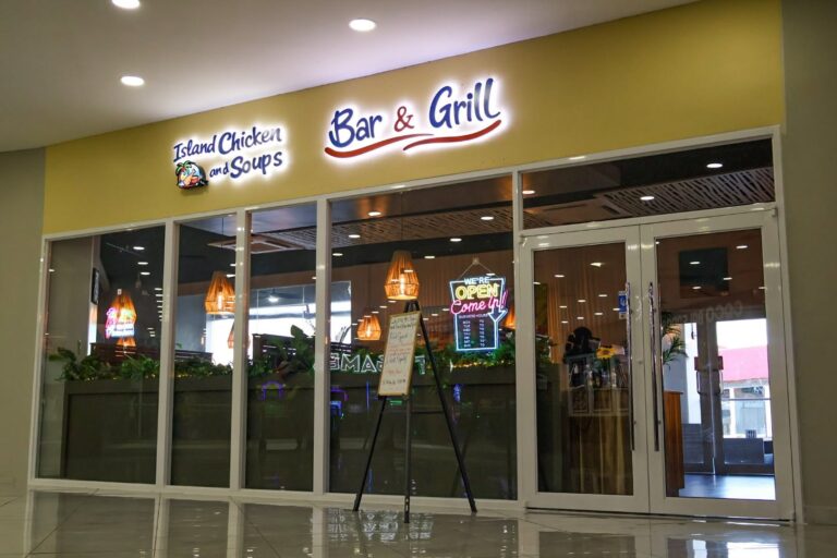 Exterior view of Island Chicken and Soups Bar & Grill located in Roatan, Honduras, showing its glass entrance doors, neon signs, and hanging wooden light fixtures inside the restaurant.