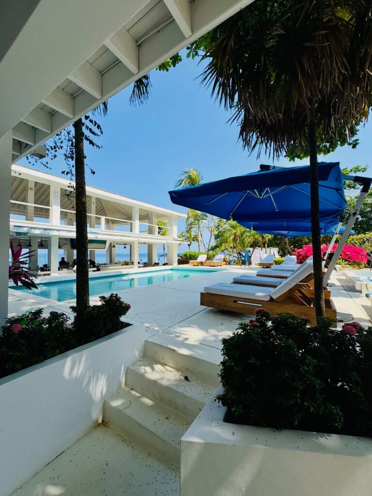 View of a luxurious swimming pool area with white lounge chairs, blue umbrellas, and tropical plants under a partly shaded building in Roatan, Honduras.