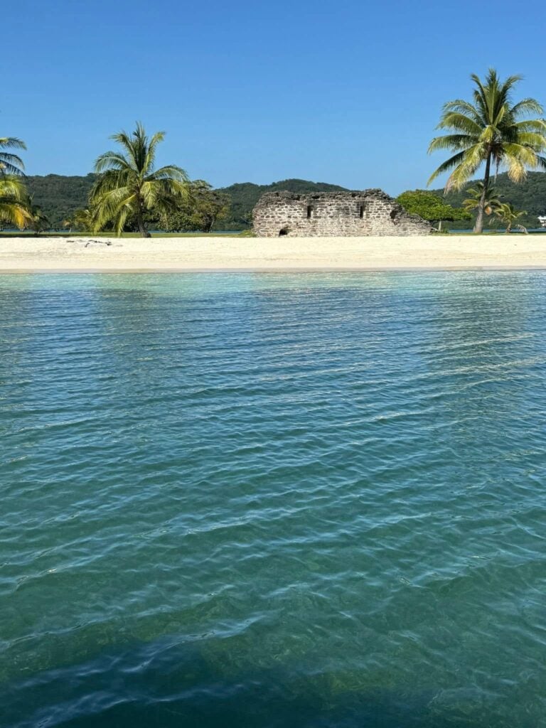 Clear turquoise waters surround a sandy beach with palm trees and ancient stone ruins on Roatan, Honduras, with lush green hills in the background.