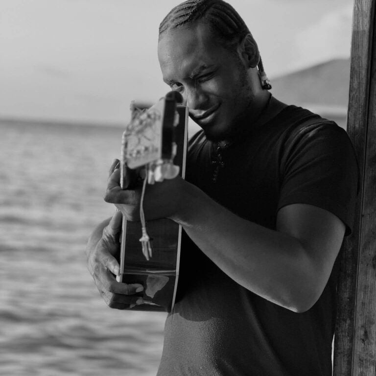 A black and white image of a woman with braided hair playing an acoustic guitar near the ocean in Roatan, Honduras, smiling and winking at the camera.
