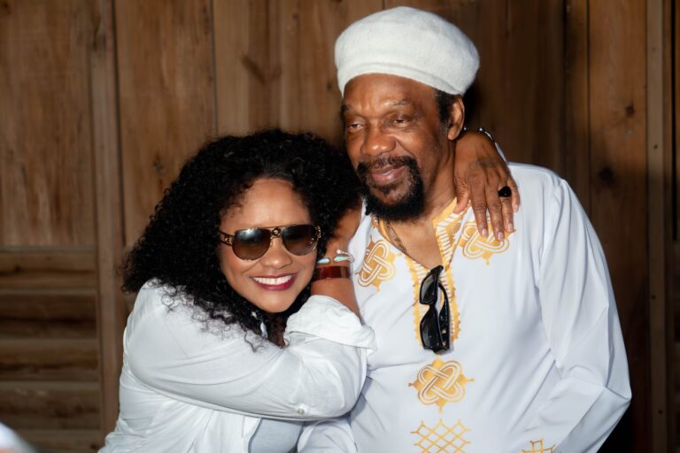 A joyful African-American couple dressed in white embracing each other in front of a wooden wall on Roatan, Bay Islands, Honduras, with the woman wearing sunglasses and smiling.