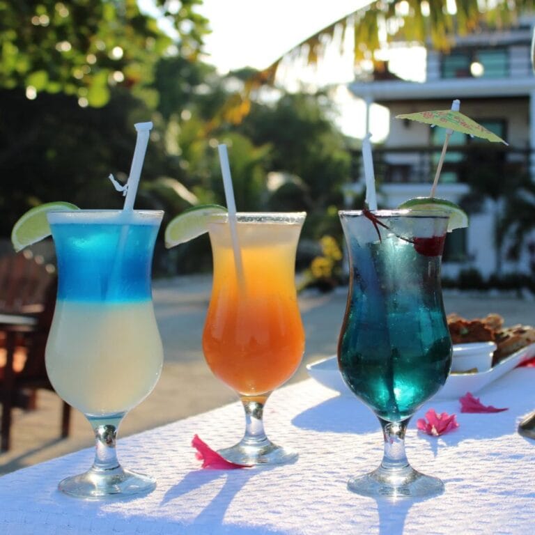 Four colorful tropical cocktails with lime wedges and tiny umbrellas are displayed on a white tablecloth against a backdrop of lush greenery and a building on Roatan, Honduras.