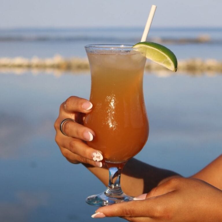 A person holds a tropical cocktail with a lime wedge and straw in front of Roatan’s beachfront with calm sea waters and a distant rocky shoreline in Honduras.