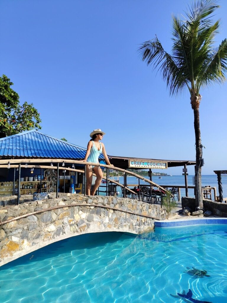 A woman in a striped swimsuit and wide-brimmed hat stands on a balcony overlooking a swimming pool with clear blue water, palm trees, and a sign for a craft cooking bar and restaurant in Roatan, Honduras.