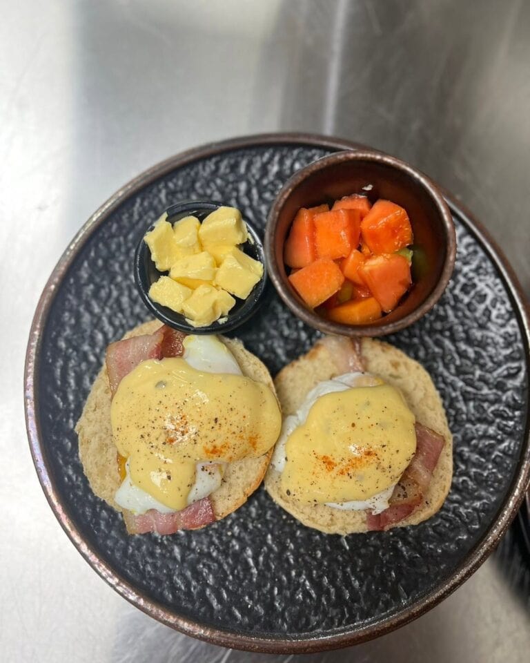 Two toasted English muffins topped with poached eggs, bacon, and hollandaise sauce, served with diced pineapple and a small bowl of chopped tomato salsa on a textured black plate in Roatan, Honduras.