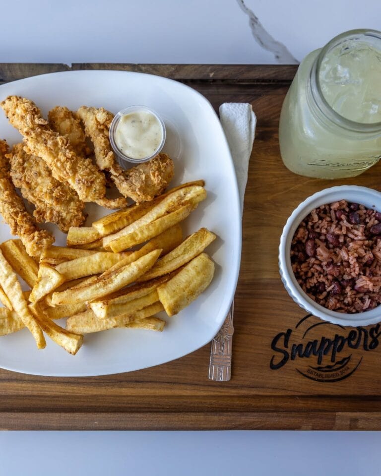 A plate of crispy fried chicken tenders with French fries and a small cup of dipping sauce sits on a wooden table in Roatan, Honduras, accompanied by a jar of lemonade and a bowl of seasoned rice.