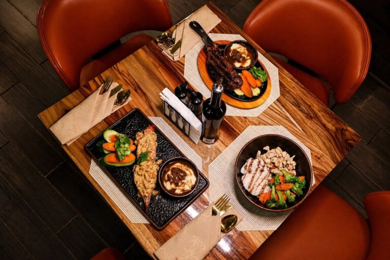 A wooden dining table set for three with plates of steak, vegetables, and rice in a restaurant on Roatan, Honduras, featuring warm chairs and neatly arranged napkins and cutlery.