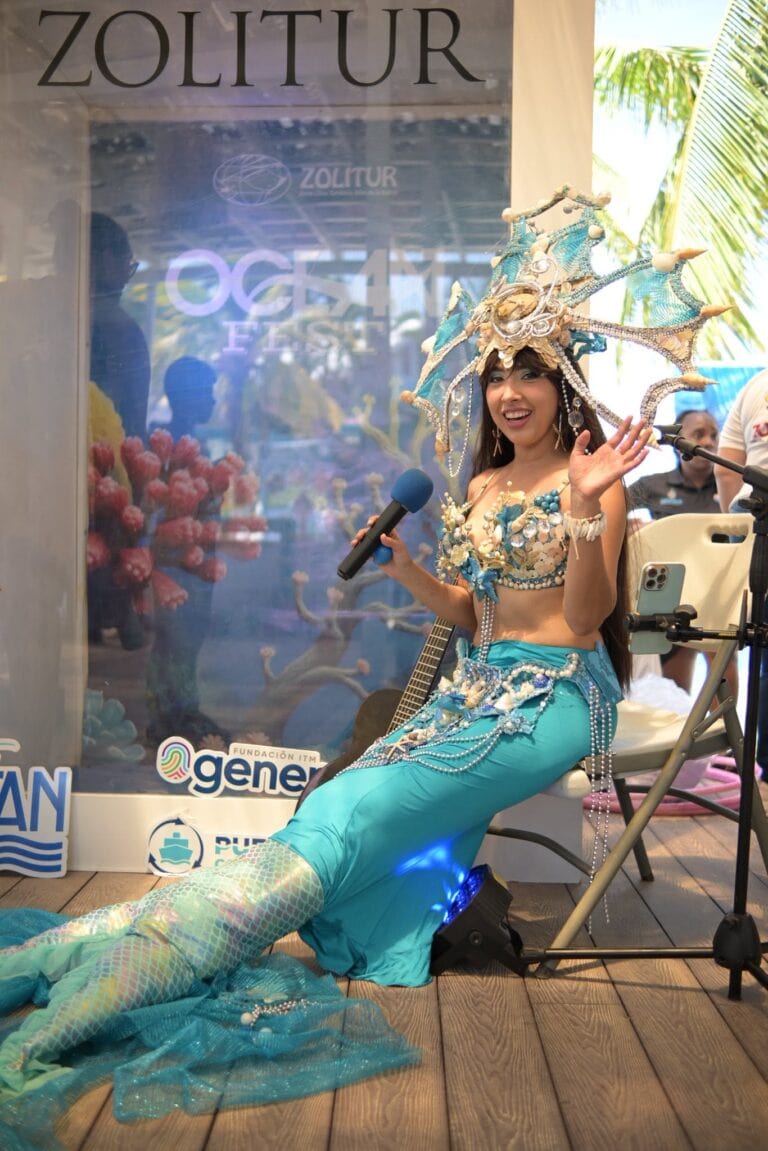A woman dressed as a mermaid with a blue tail and intricate seashell and pearl accessories, smiling and waving at an event in Roatan, Honduras, standing in front of a promo display with tropical palm trees in the background.