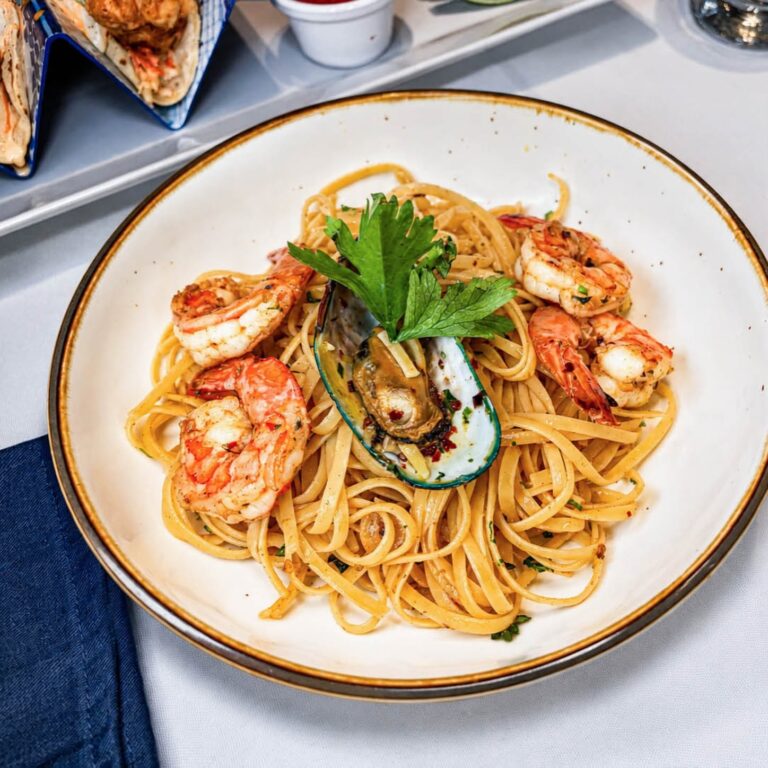 A Plate of spaghetti pasta topped with large shrimp, an open oyster, and fresh herbs served on a white dish with a decorative border, with partially visible tacos and condiments in the background on a table in Roatan, Honduras.