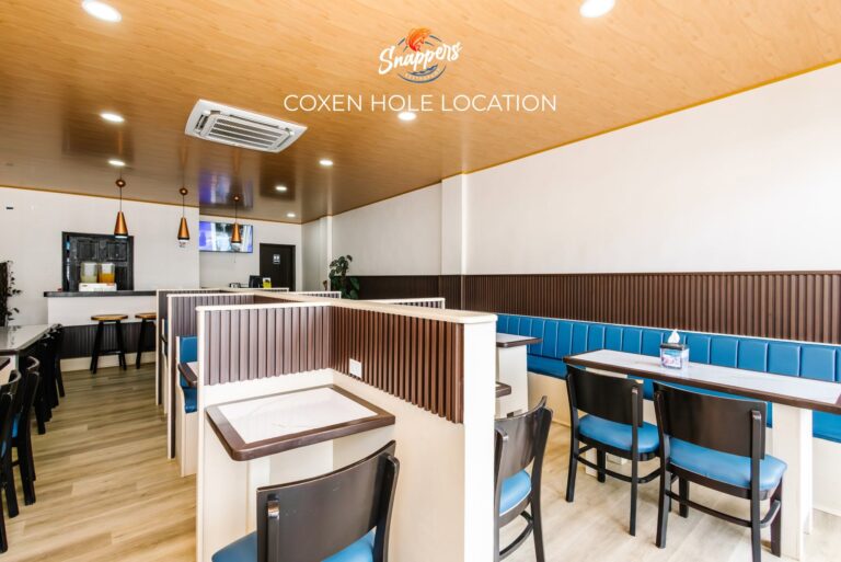Interior view of a modern restaurant in Roatan, Honduras, featuring a light wood ceiling, white walls, blue cushioned seating along the wall, and black tables with black and blue chairs, decorated with small potted plants and a wall-mounted television.