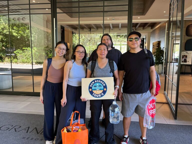 Group of five diverse tourists standing at Roatan's airport with luggage and tour sign in front of glass doors showing greenery outside in Honduras.
