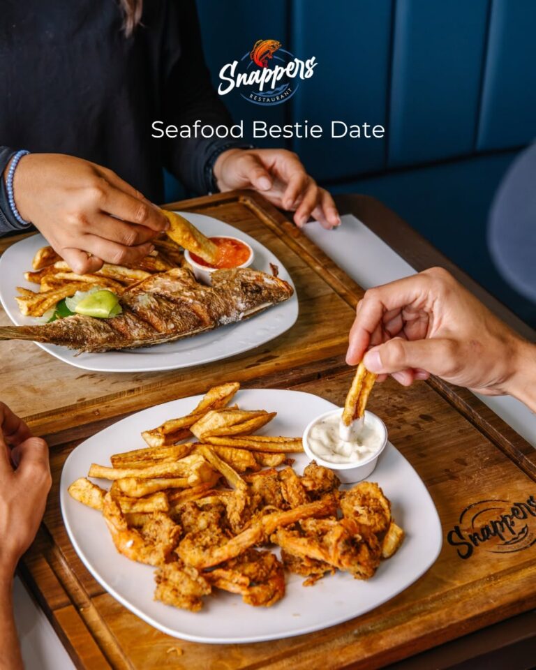 Two people enjoying a seafood meal at Snappers Restaurant in Roatan, Honduras, with a grilled fish, fried seafood, and sides on wooden trays.