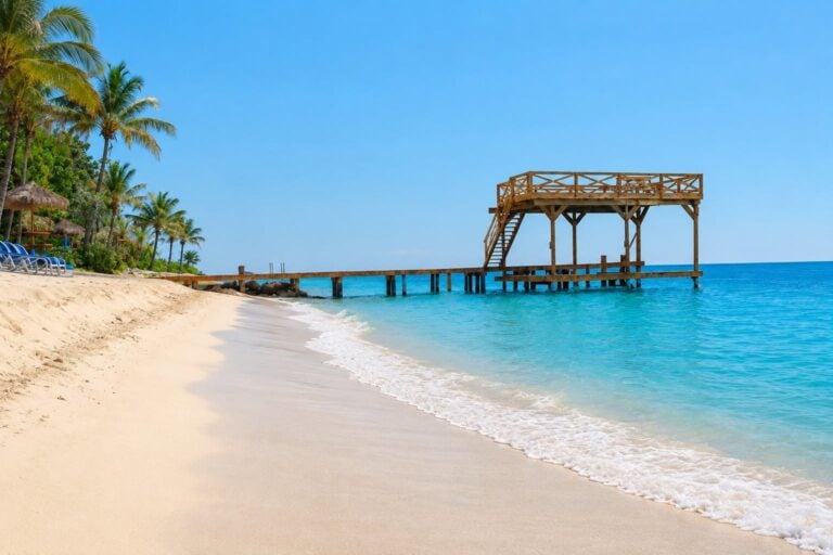A wooden pier with a raised observation deck extends into the clear turquoise waters of Roatan, Honduras, with a sandy beach and lush palm trees along the shoreline under a bright blue sky.