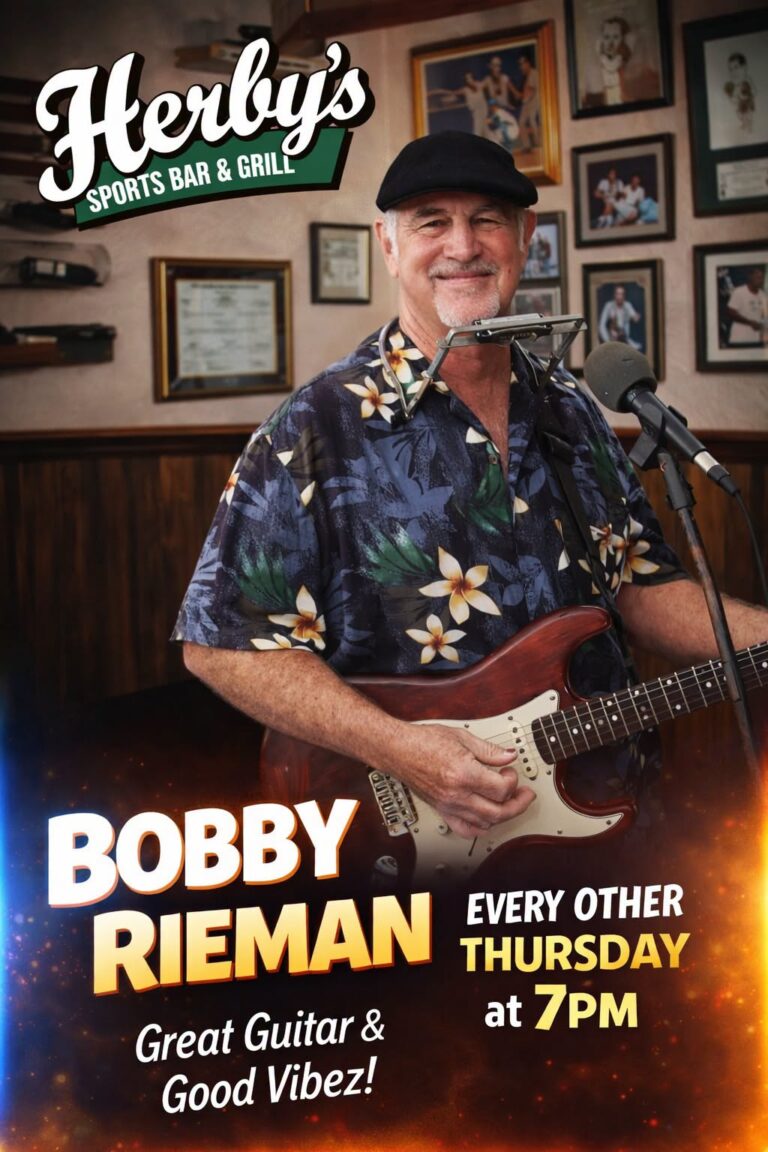 Bobby Rieman performing live music at Herby’s Sports Bar & Grill in Roatan, Honduras with a guitar and harmonica in a cozy indoor setting decorated with framed photographs on the wall.