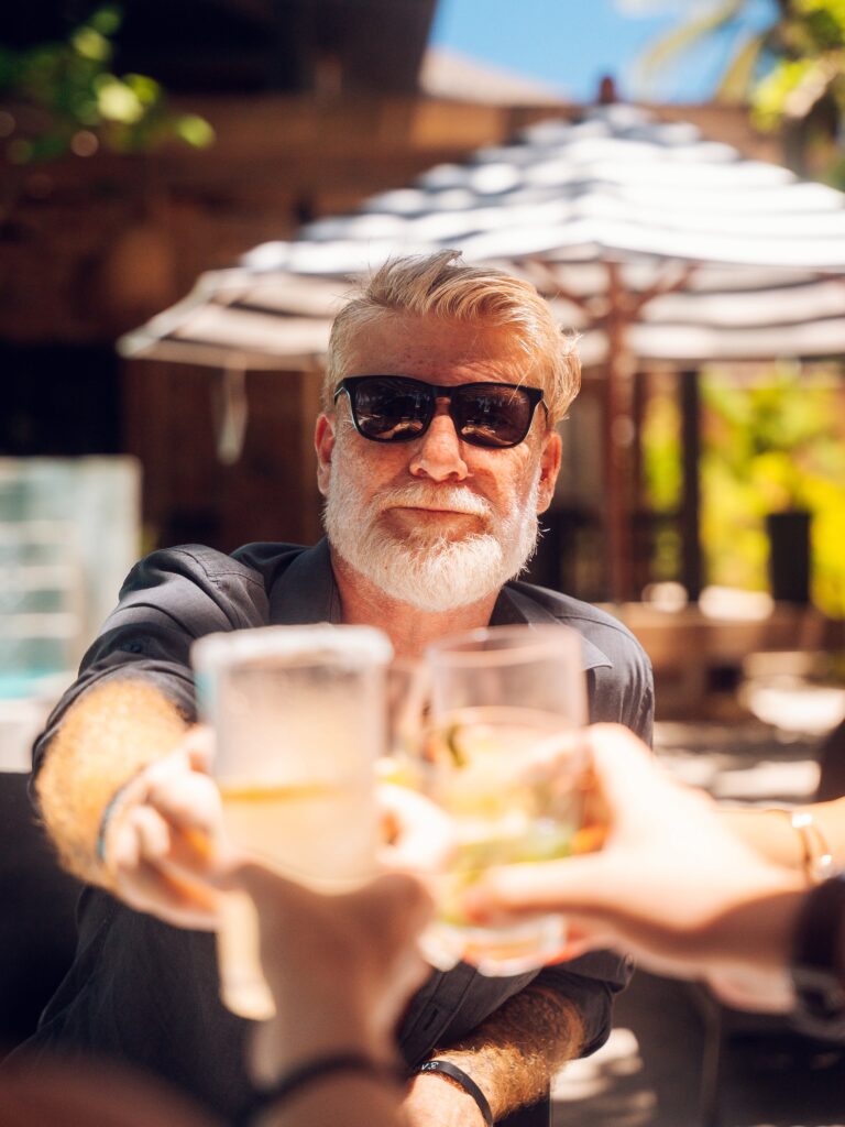 Close-up of a smiling elderly man with white hair and beard wearing dark sunglasses at an outdoor table in Roatan, Honduras, with a blurred background of a white and brown striped patio umbrella and greenery.
