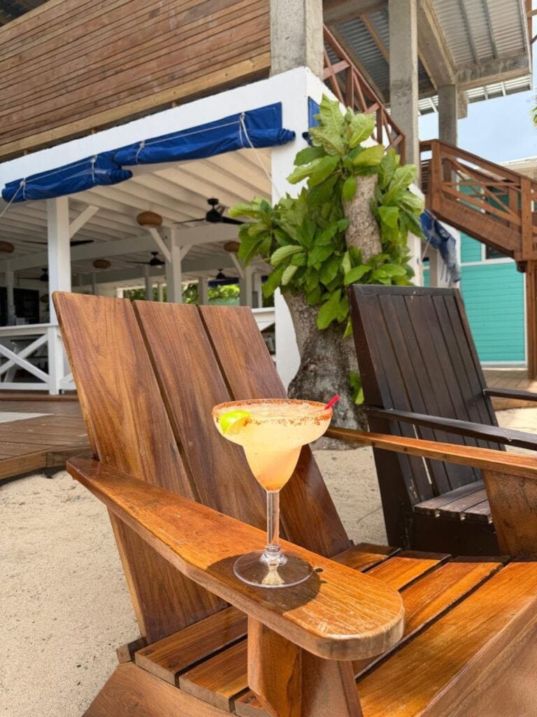 A mango margarita with a lime wedge on a wooden lounge chair at a beachfront bar in Roatan, Honduras, with a tropical setting including a tree and a beachside building in the background.
