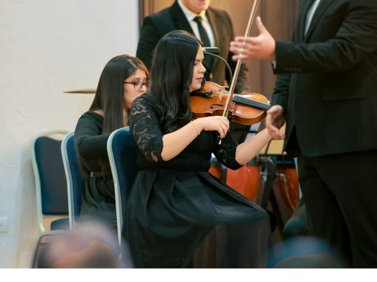A woman playing the violin during a formal event or concert in Roatan, Honduras, with musicians and a conductor visible in the background.