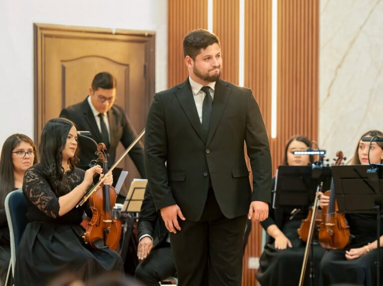 A man in a black suit and tie stands on stage with an orchestra in Roatan, Honduras, during a formal musical performance.