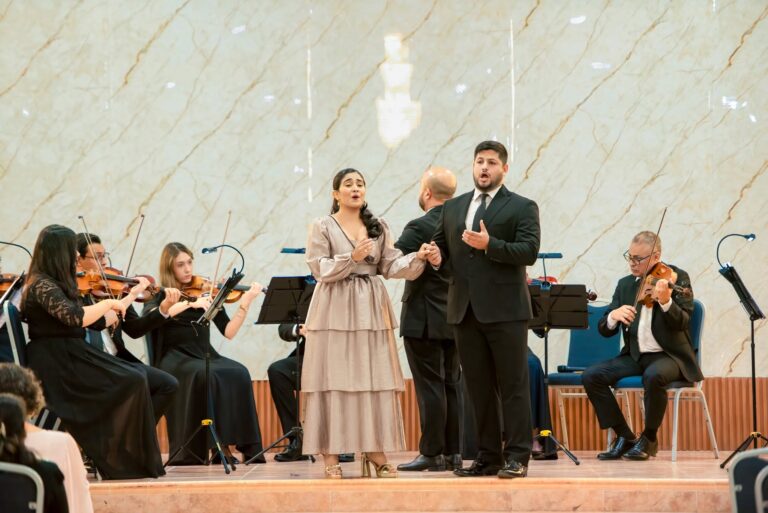 Opera singers perform passionately on stage in a concert hall in Roatan, Honduras, surrounded by an orchestra of string players and a marble wall backdrop.