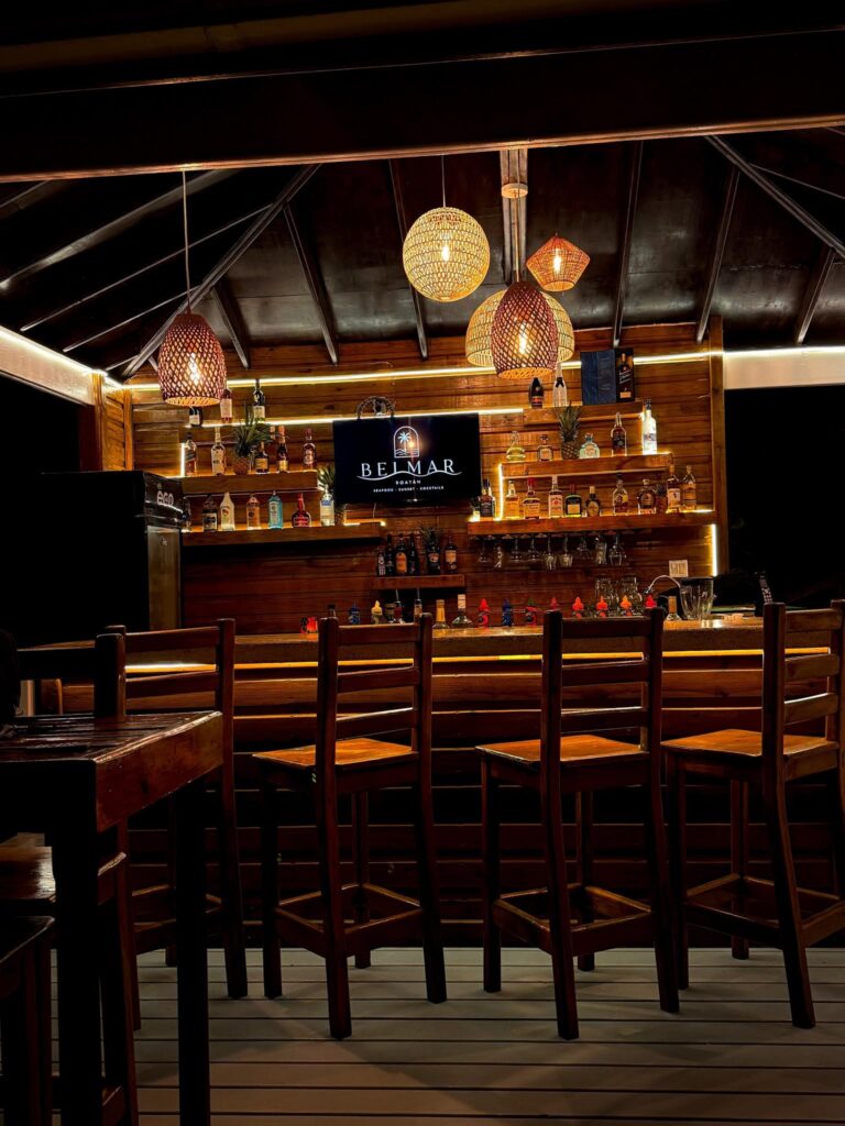 Rustic wooden bar interior in Roatan, Honduras, featuring hanging woven lanterns, liquor shelves, and rows of tall bar stools.