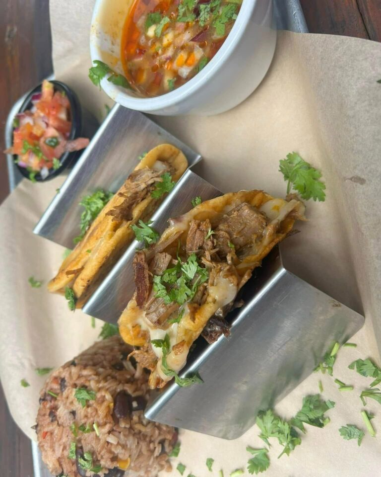 Three tacos filled with shredded meat, cheese, and vegetables served on a metal tray with a side of rice and beans in a restaurant setting on Roatan, Honduras.