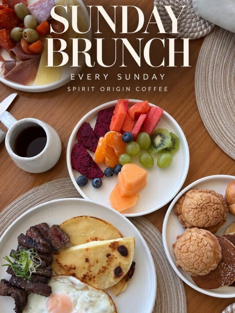 An assortment of breakfast dishes including fresh fruit, eggs, toast, and baked goods arranged on a wooden table in Roatan, Honduras, with text promoting Sunday brunch at Spirit Origin Coffee.