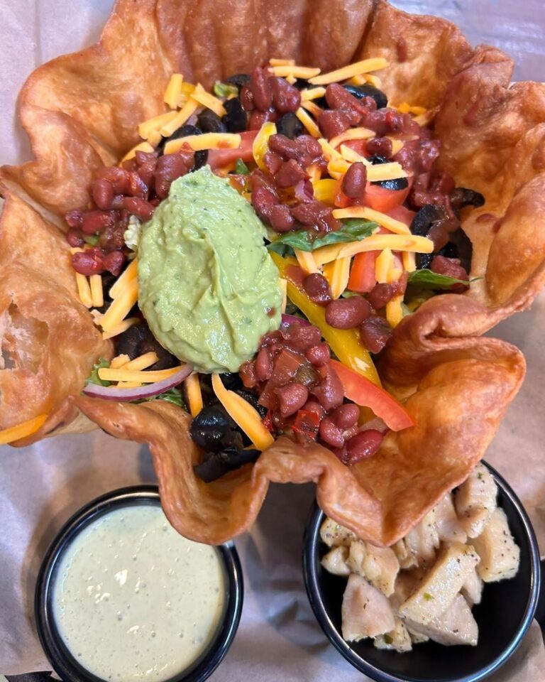 A crispy tortilla bowl filled with black beans, shredded cheddar cheese, fresh tomato slices, chopped red onion, and a dollop of guacamole, set on a wooden table in Roatan, Honduras.