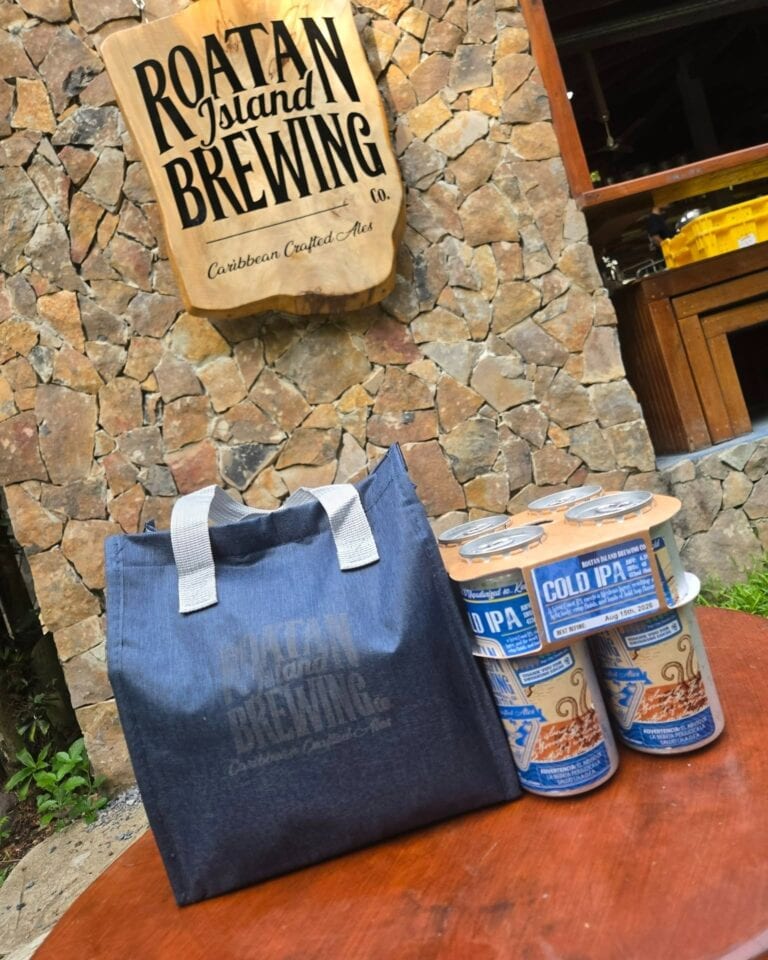 Wooden sign reading 'Roatan Island Brewing Co.' mounted on a stone wall inside a brewery in Roatan, Honduras, with blue tote bag and several cans of Cold IPA beer on a wooden table.