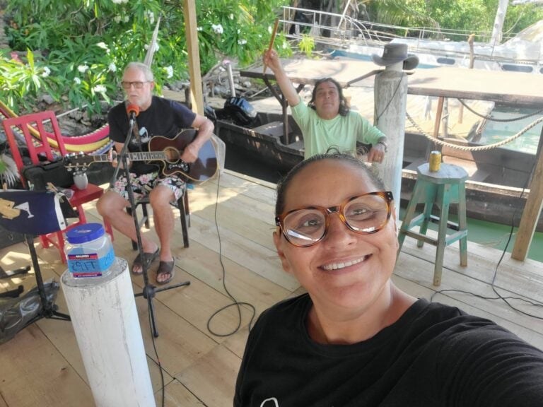 Two musicians perform on a wooden deck in Roatan, Honduras, with lush green tropical foliage and a docked boat in the background.