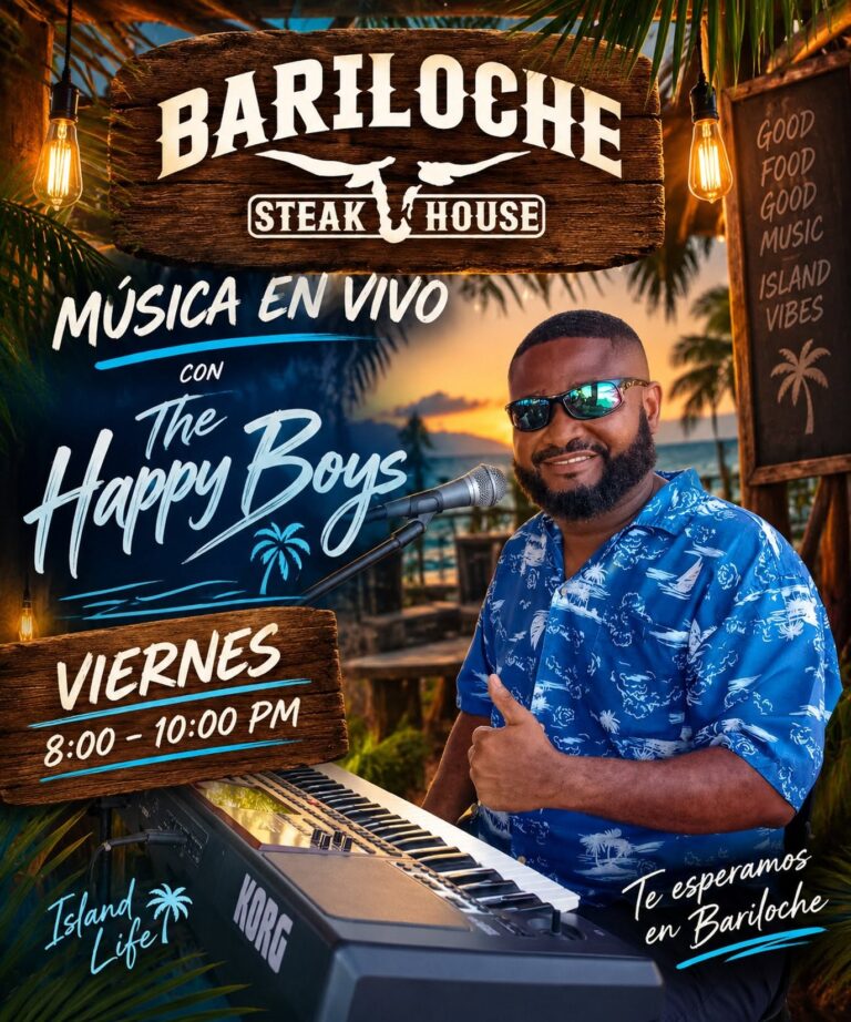 A man with a beard and sunglasses dressed in a blue Hawaiian shirt smiling next to a keyboard at a live music event in Roatan, Honduras, with palm trees and a sunset in the background.