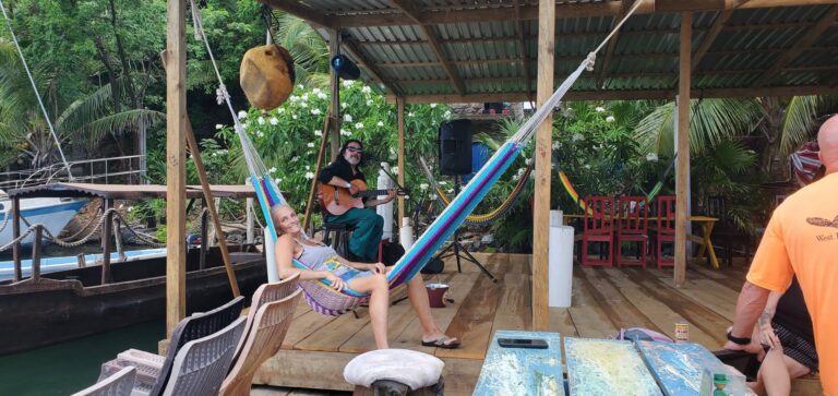 Musicians performing live on a wooden deck at a tropical outdoor venue in Roatan, Honduras, with lush green foliage and flowering plants in the background.