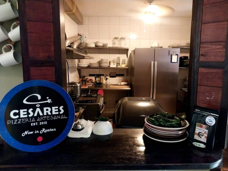 Indoor view of Cesáres Pizzeria Artesanal in Roatan, Honduras, displaying a kitchen with shelves of plates, glasses, condiments, and cooking utensils behind a dark wooden counter.