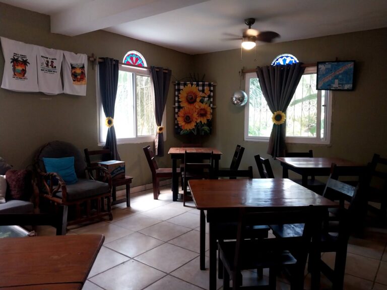 Interior of a cozy dining area in Roatan, Honduras, with wooden tables and chairs, sunflowers on the curtains and wall decorations, and a small TV screen showing a map of Roatan Island.