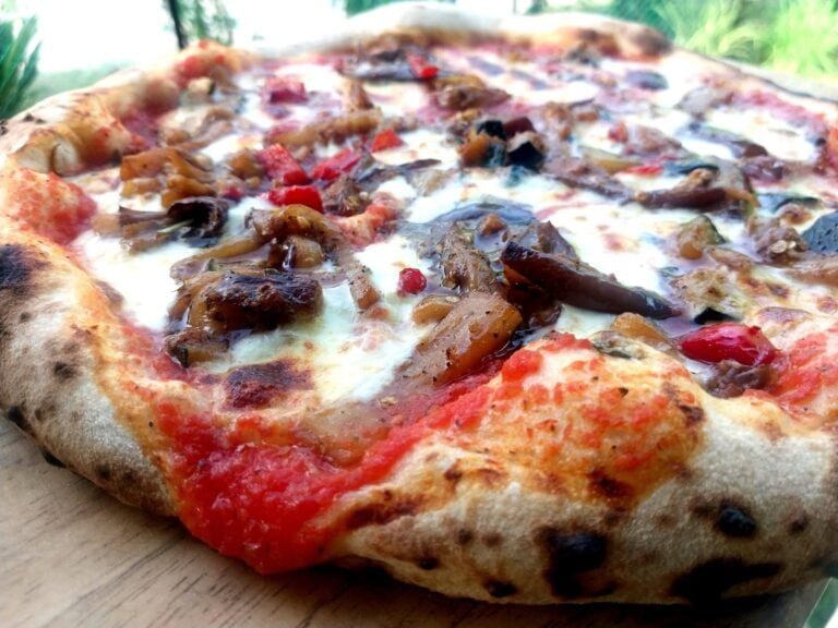 Close-up of a freshly baked pizza topped with mushrooms, red peppers, and melted cheese on a wooden surface, set against a blurred outdoor background in Roatan, Honduras.