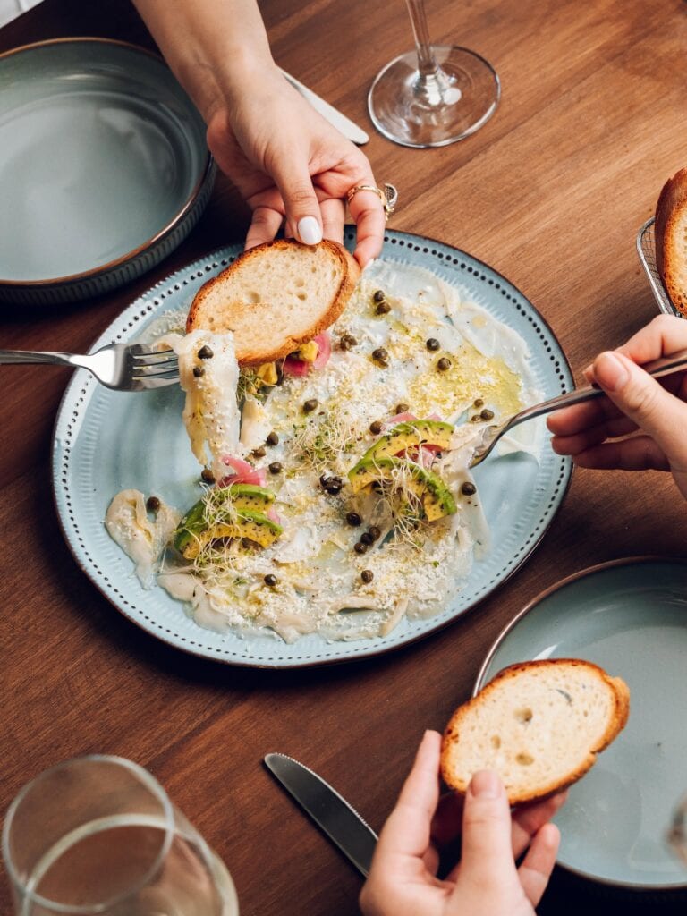 A plated seafood dish with herbs, capers, and toasted bread is shared at a restaurant table in Roatan, Honduras.