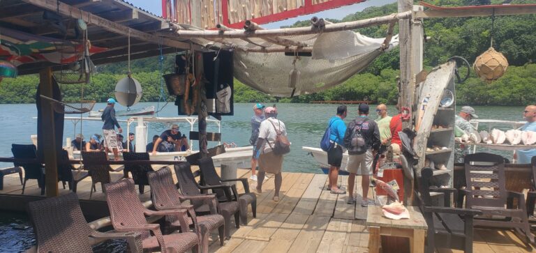 A busy dock in Roatan, Bay Islands, Honduras, with tourists boarding a small boat surrounded by lush green hills and calm water.