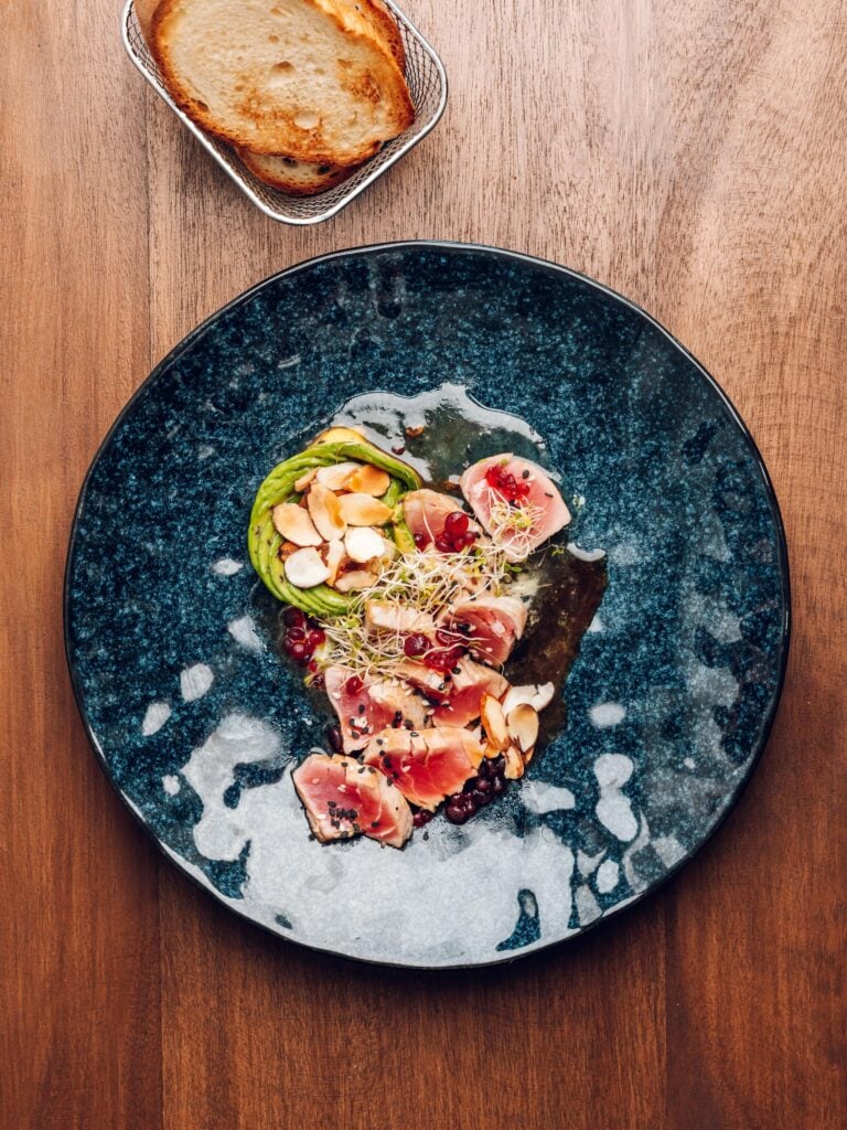 A gourmet plated dish with sliced vegetables, herbs, edible flowers, and toasted bread on a blue ceramic plate in Roatan, Honduras.