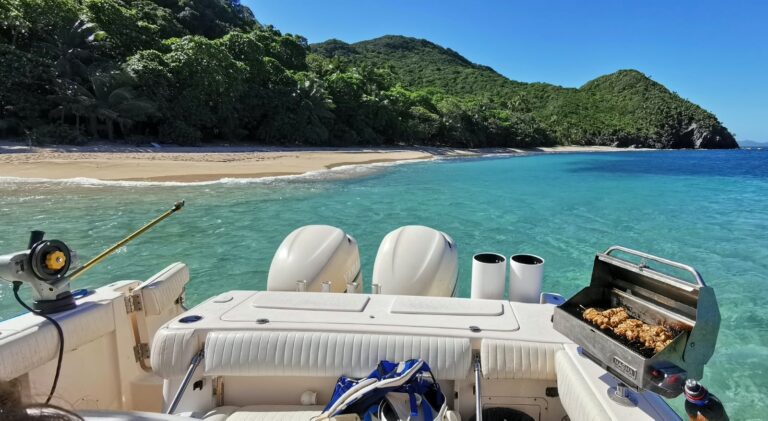 A motorboat with twin outboard engines and an open grill floats in turquoise water beside a sandy beach and lush hills in Roatan, Honduras.