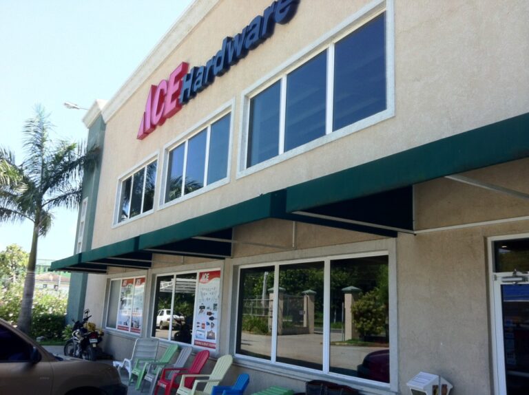 A two-story Ace Hardware store in Roatan, Honduras, with large windows, a green awning, colorful outdoor seating, and palm trees in the background.